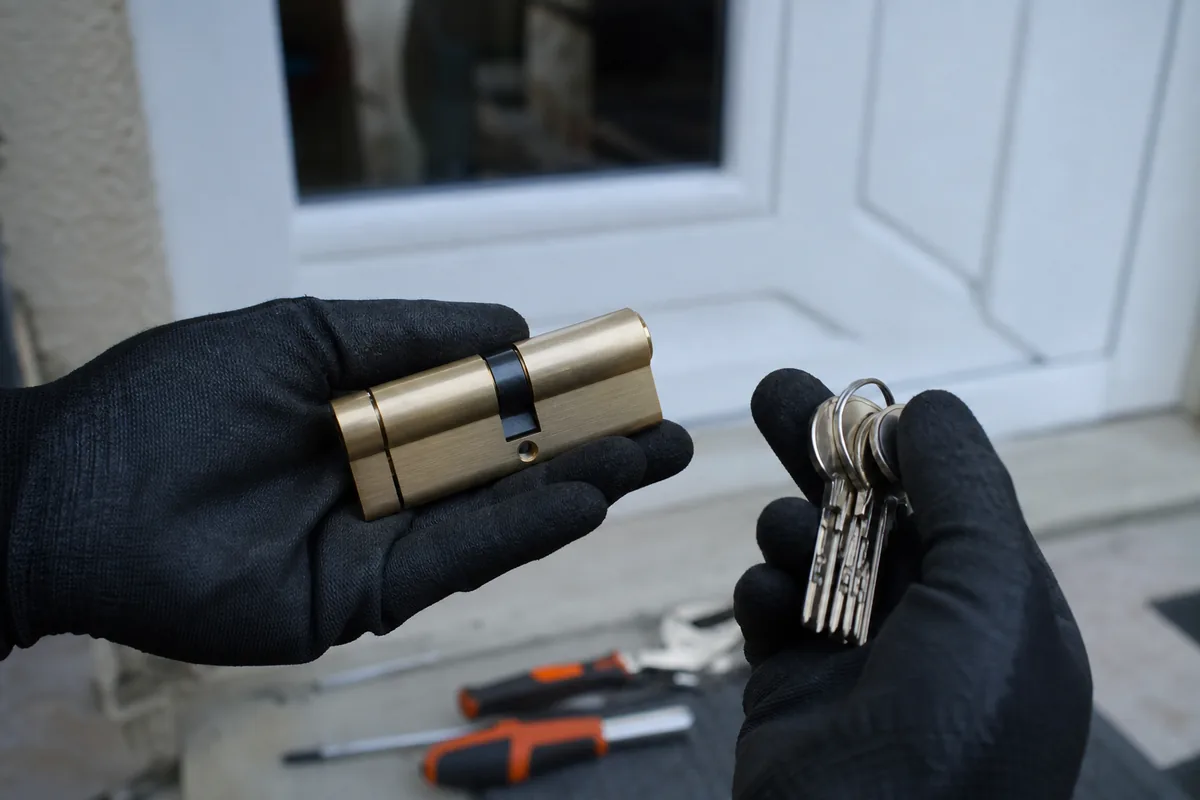 Mains gantées d’un serrurier tenant un cylindre européen et un trousseau devant une porte d’entrée